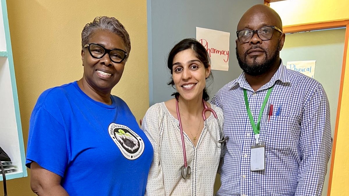 Three Impact Life team members standing together inside the Bunkers Hill Community Health Center — one in a blue Impact Life International t-shirt, one with a stethoscope, and one wearing a volunteer badge, with a handwritten Pharmacy sign on the wall behind them