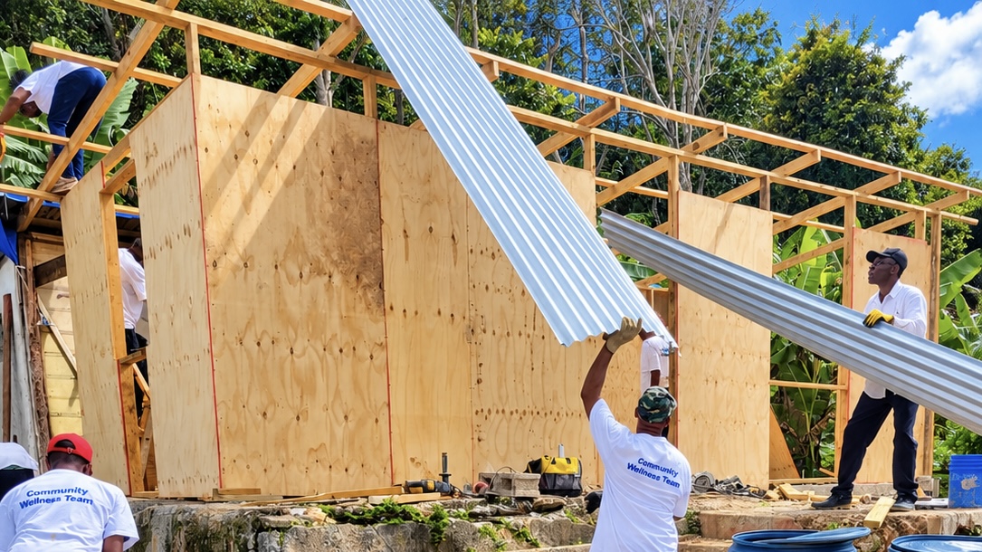 Impact Life volunteers in white Community Wellness Team shirts raising a corrugated roofing sheet onto the timber frame of a building under construction in Jamaica
